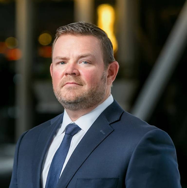 Professional headshot of Brett Cain, Texas personal injury attorney, in navy suit and tie against blurred office background.
